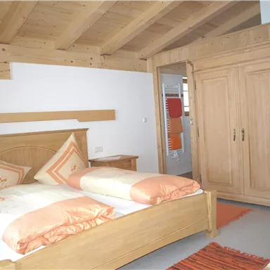 A cozy room with a wooden bed and orange decorative pillows. The room has a light wooden ceiling and a large wardrobe.
