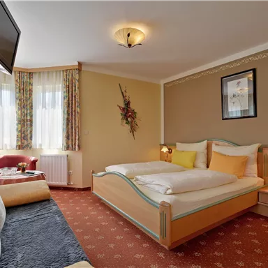 A cozy bedroom with a double bed and colorful pillows. There is a sofa, a TV, and large windows that let in plenty of light.