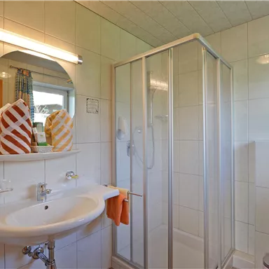 A modern bathroom with a shower cabin, a sink, and a large mirror. The walls are tiled, and there are fresh towels.