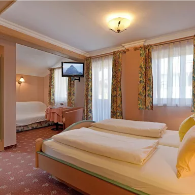 A cozy hotel room with two beds and a TV. There are large windows with curtains and a separate sleeping area.