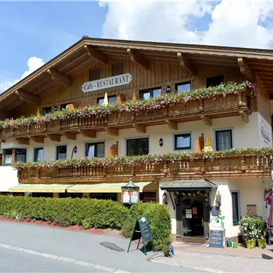 A charming building with a restaurant sign and many blooming balcony boxes. The sky is blue and there is a green surrounding.