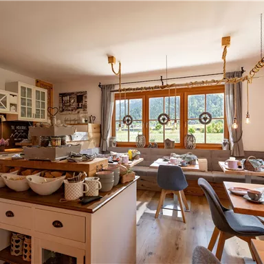 A cozy breakfast room with wooden furniture and large windows. Various foods and drinks are arranged on the table.