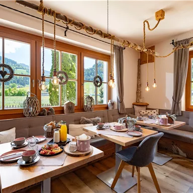 An inviting breakfast room with wooden furniture and large windows. The table is lovingly set with food and drinks and offers a view of the mountains.