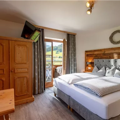 A cozy bedroom with a large bed and wooden furniture. The room has a balcony overlooking the landscape.