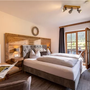 A cozy bedroom with a large bed and wooden accents. The windows offer a view of nature and let in plenty of light.
