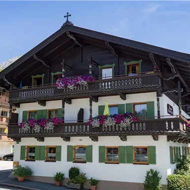 A traditional alpine wooden house with green shutters and colorful flowers on the balcony. In the background, the mountains and a clear blue sky can be seen.