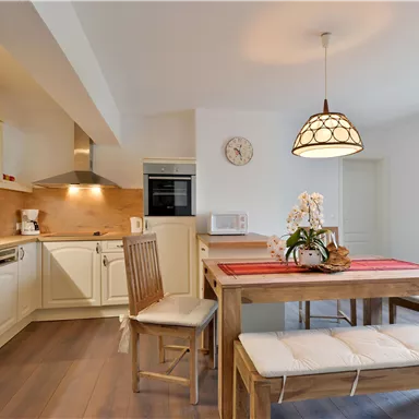 A bright kitchen with modern furniture and a wooden dining table. The walls are neutral and there are colorful curtains at a window.