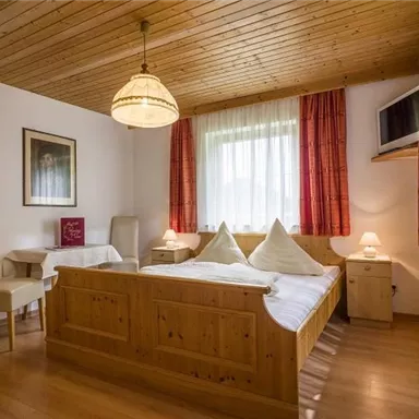 A cozy bedroom with a wooden bed and two pillows. The walls are light, and the room has a warm atmosphere with windows and curtains.
