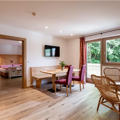 A cozy living room with a dining area, chairs, and a television. Large windows offer a view of the nature outside.