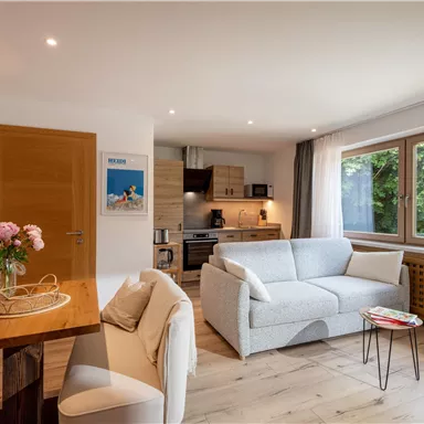 A bright and modern living kitchen with a cozy sofa and a dining table. Large windows offer a view of the green surroundings.