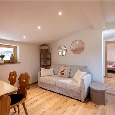 A bright, modern living room with a cozy sofa and a dining table. The room has large windows and friendly, neutral colors.