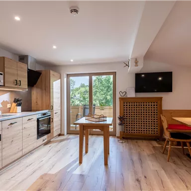 A modern kitchen with wooden cabinets, stove, and microwave. The dining area is cozy with a red bench and a table.