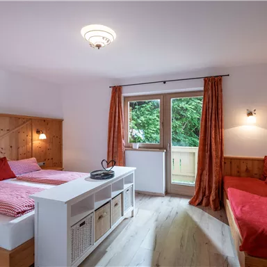 A cozy bedroom with wooden furniture and red accents. The room features a window with a view of greenery and an inviting seating area.