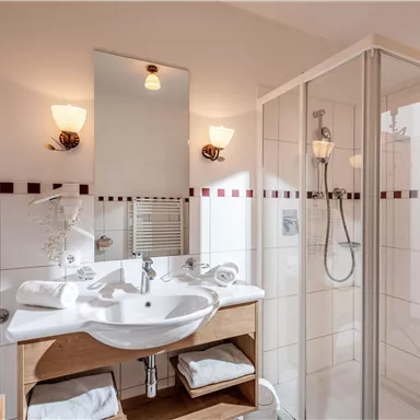 A modern bathroom with a shower and a sink. The walls are light-tiled and there are towels on a shelf.