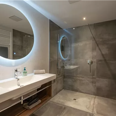 A modern bathroom with a large mirror and a walk-in shower. The walls are in a simple gray tone, with elegant fixtures and towels.