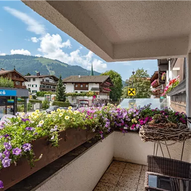 A balcony with colorful flowers and a view of the mountains. In the background, houses and a clear sky can be seen.
