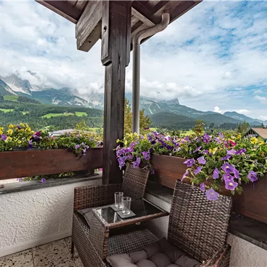 A cozy balcony with a table and chairs, surrounded by colorful flowers. In the background, impressive mountains and a clear sky can be seen.