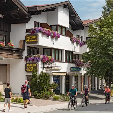 A picturesque street with traditional buildings and colorful flower boxes. People walk and ride bicycles past, while trees provide shade.