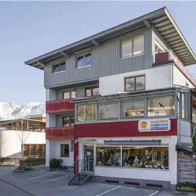 A modern building with several floors and large windows. In the background, mountains and a clear sky are visible.