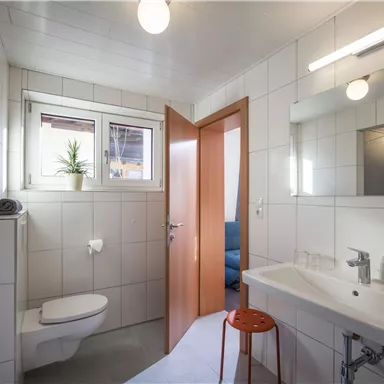 A modern bathroom with white tiles and wooden details. It includes a toilet, a sink, and a large mirror.