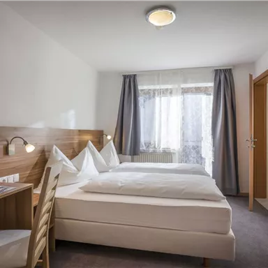 A modern hotel room with two beds and a desk. The windows let in a lot of light and the walls are painted in neutral tones.