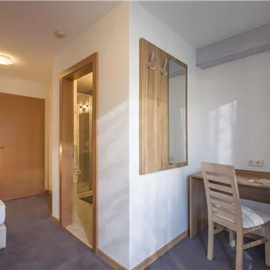 A modern hotel room with a bed, a desk, and an adjoining bathroom. The walls are painted in light tones, and there is plenty of daylight.