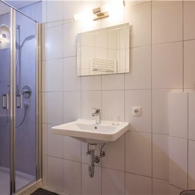 A modern bathroom with a shower and a sink. The walls are equipped with white tiles and there is a large window for natural light.
