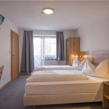 A modern hotel room with two single beds and a television on the wall. Large windows provide plenty of daylight.