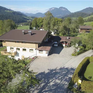 Ein charmantes Haus umgeben von grünen Hügeln und Bäumen. Im Hintergrund sind Berge sichtbar, die eine malerische Landschaft bilden.