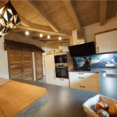 A modern kitchen with wooden ceilings and elegant lamps. In the background, an impressive mountain landscape can be seen.