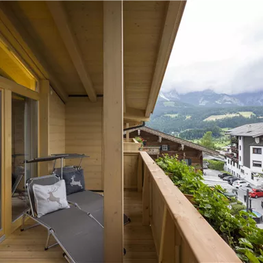 Ein Balkon mit Blick auf die Berge und die umliegenden Gebäude. Die Holzverkleidung und die gemütliche Liege laden zum Entspannen ein.