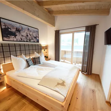 A modern bedroom with a large double bed and wooden details. Large windows provide plenty of daylight and a view outside.