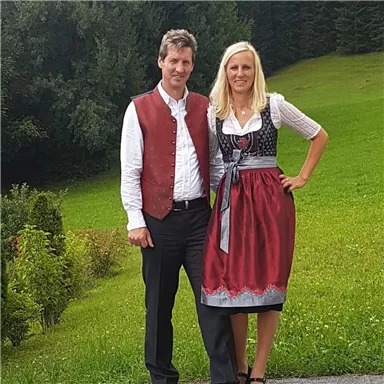 Ein Paar in traditioneller bayerischer Tracht steht vor einer grünen Wiese. Die Frau trägt ein elegantes Dirndl und der Mann eine Weste.