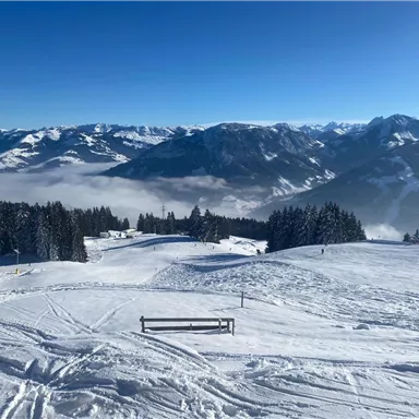 A snow-covered landscape with mountains in the background. The sky is clear and blue, while fog is visible in the valley.