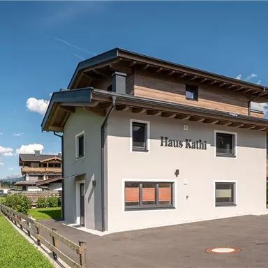 A modern house named "Haus Kathl" in a green environment. The facade is bright and friendly, surrounded by meadows and mountains.