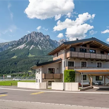 A modern building in the midst of a picturesque mountain landscape. The surroundings are green and the clouds above the mountains are clearly visible.