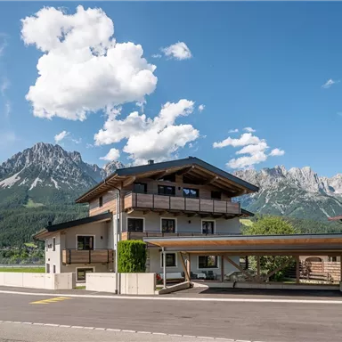 A modern house with a wooden carport in front of a picturesque mountain landscape. The sky is blue with some clouds.