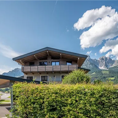 A modern building with a curved roof and large windows. In the background, majestic mountains and a clear blue sky can be seen.