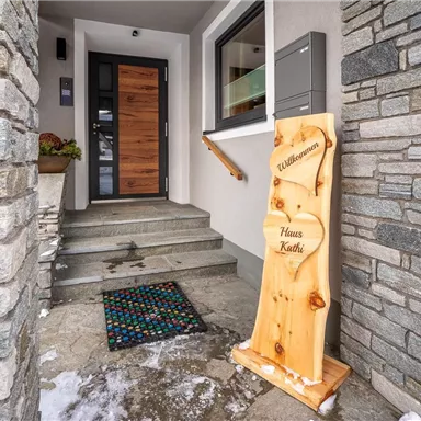 Entrance to a modern house with a wooden door and a welcoming doormat. Next to the door is a welcome sign with the inscription "House Kathi".