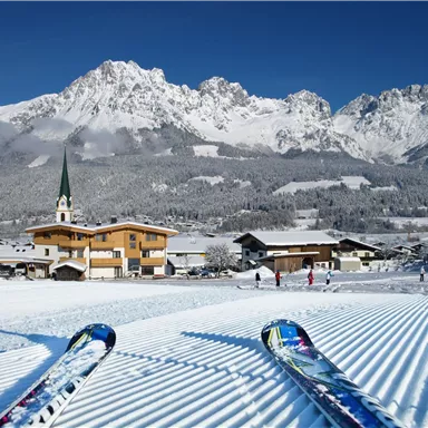 A picturesque winter scene with snow-covered mountains and a small village. In the foreground, skis are on freshly groomed slopes.