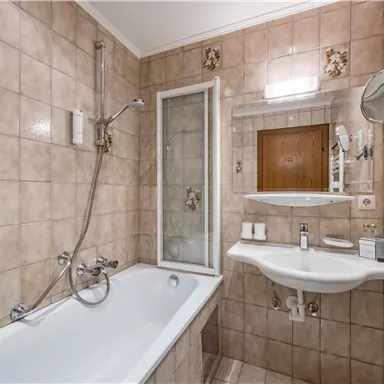 A bathroom with a bathtub and a shower cabin. The walls are decorated with light tiles and there is a sink with a mirror.