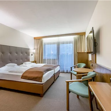 A modern hotel room with a large bed, a television, and a small table. Daylight is coming through the windows, and there are two chairs next to the table.