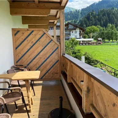 A wooden balcony with two chairs and a table. In the background, there is a green meadow and mountains.