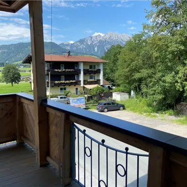 View from a balcony overlooking a green landscape with mountains in the background. In the foreground, there is a wooden railing and a building is visible.