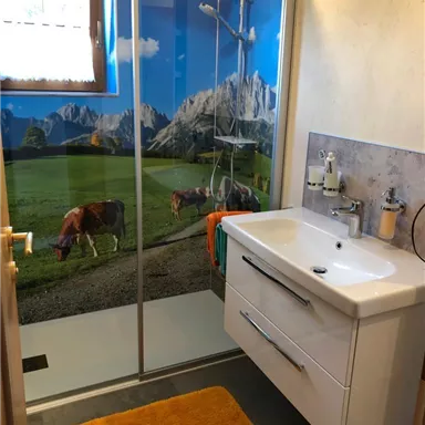 A modern bathroom with a shower and a sink. The shower wall depicts a picturesque mountain panorama with cows in a meadow.