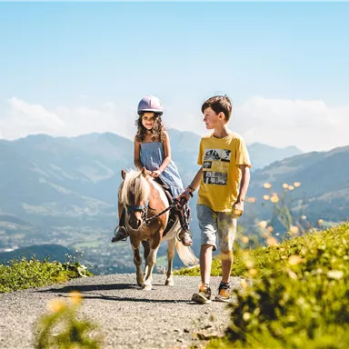 A girl is riding a pony while a boy is leading it by the reins. In the background, green mountains and a blue sky can be seen.