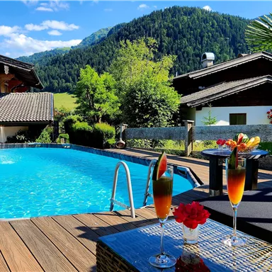 A beautiful outdoor pool surrounded by a wooden terrace and lush greenery. In the foreground are two cocktails with fresh fruits.