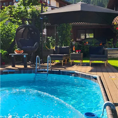 A clear pool in a beautiful garden, surrounded by plants and an attractive seating area. In the background, mountains and a blue sky can be seen.