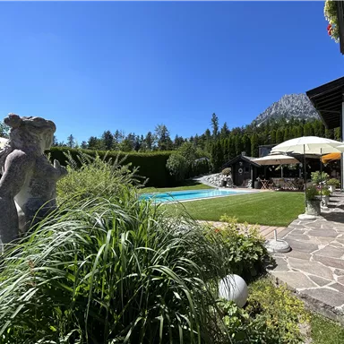 A beautiful garden with a swimming pool, surrounded by trees and green plants. A sunny sky is visible in the background.