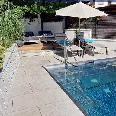 Ein moderner Garten mit einem kleinen Pool und Liegen. Neben dem Pool steht ein Sonnenschirm und es gibt grüne Pflanzen in Schalen.